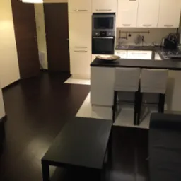 Nowoczesne wnętrze mieszkania w Trzebnicy: ciemne podłogi, białe meble kuchenne, wyspa z hokerami i kwiaty w wazonie. Minimalistyczny design i ciepłe oświetlenie.