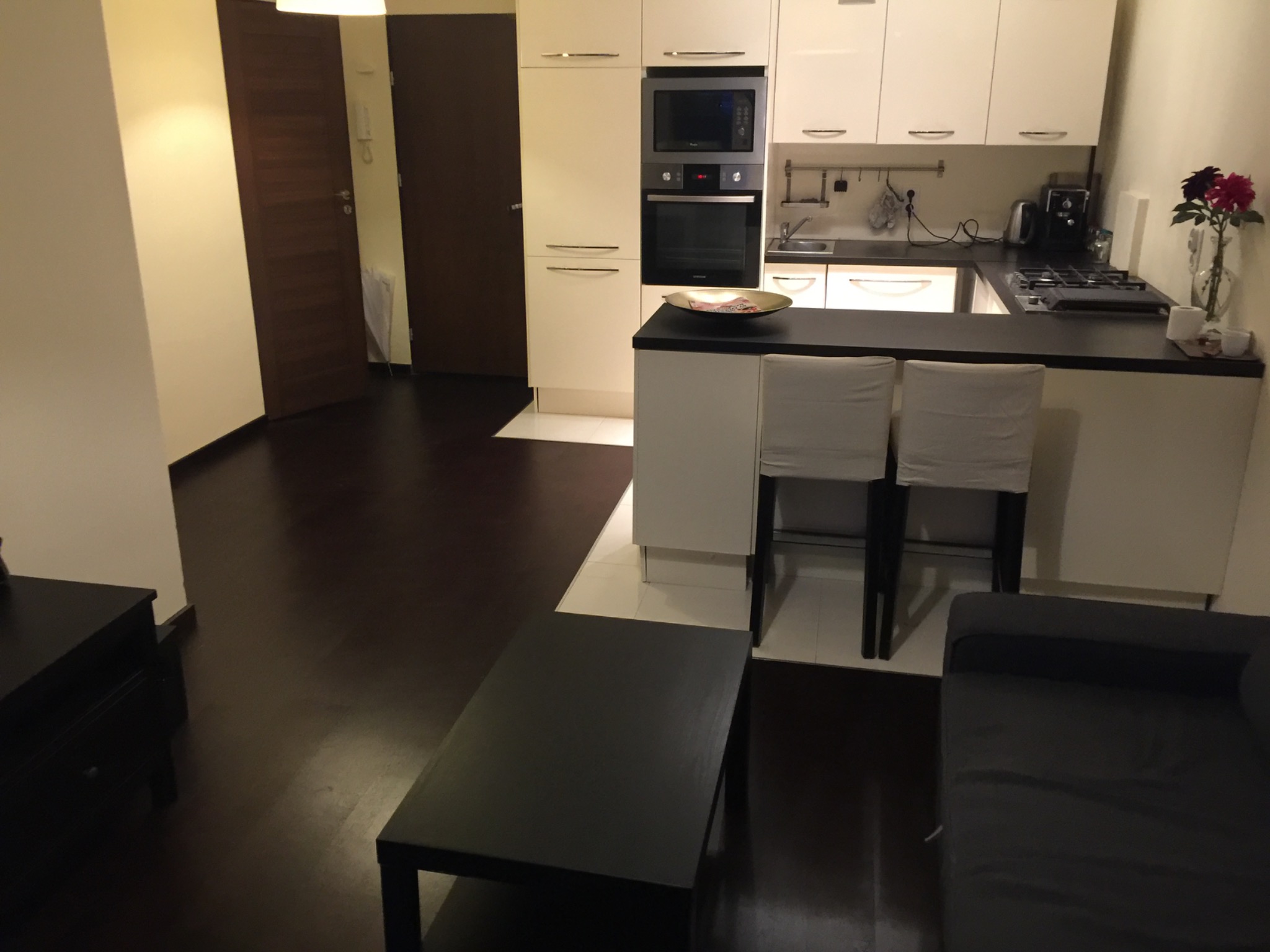 Nowoczesne wnętrze mieszkania w Trzebnicy: ciemne podłogi, białe meble kuchenne, wyspa z hokerami i kwiaty w wazonie. Minimalistyczny design i ciepłe oświetlenie.