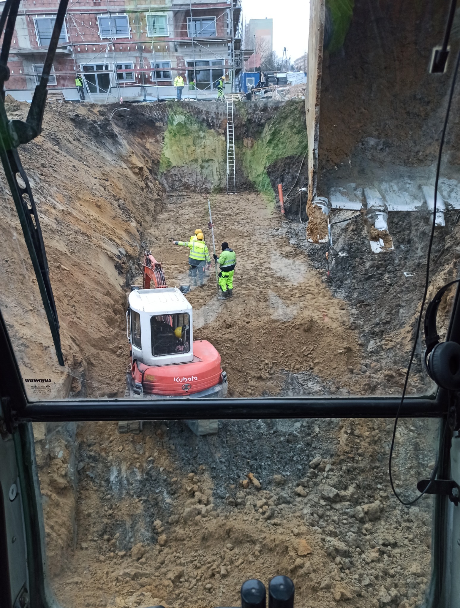 Widok z kabiny operatora na wykop z mini koparką Kubota i pracownikami w odblaskowych ubraniach; w tle budynek w rusztowaniach i drabina.