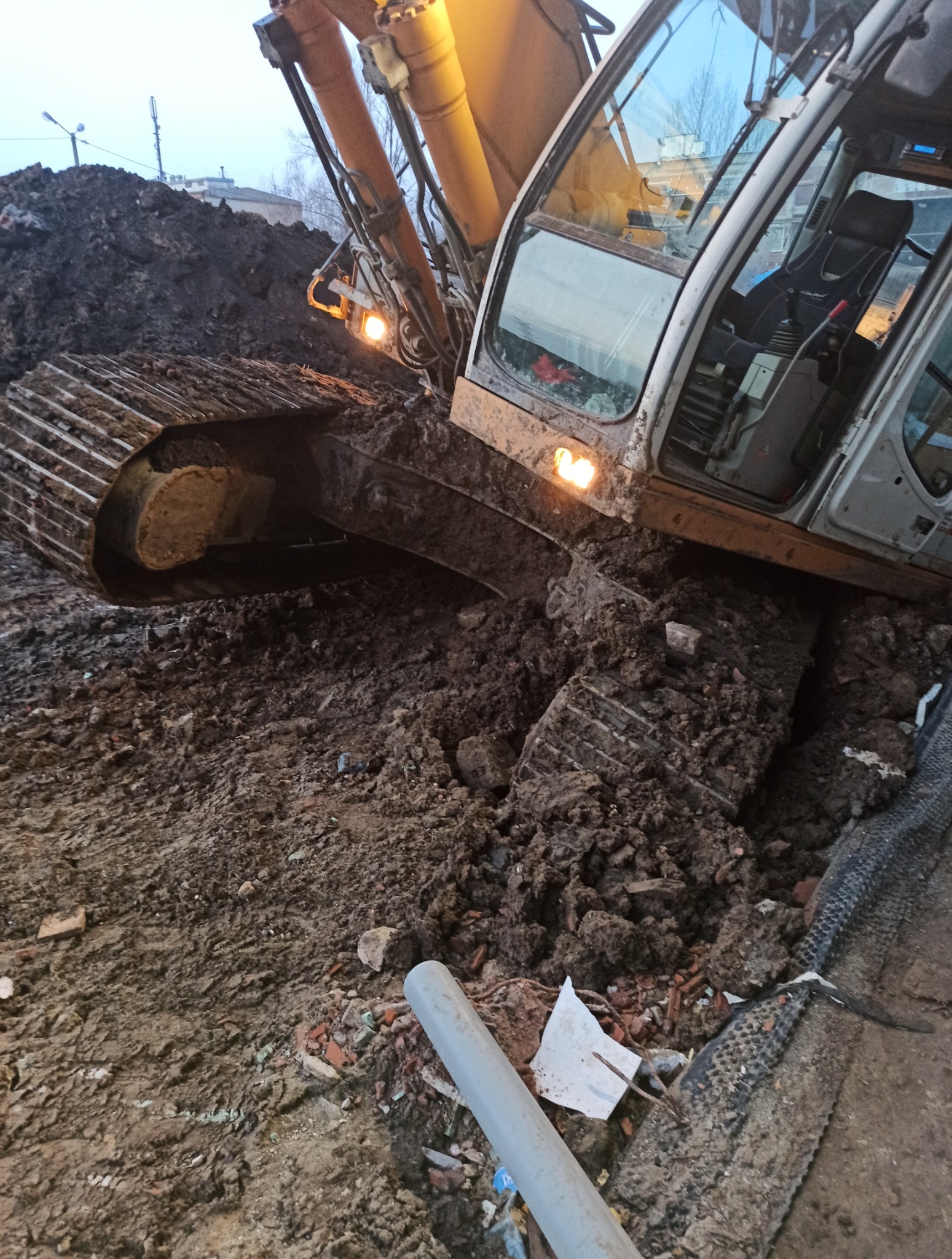 Ciężka, zabłocona koparka gąsienicowa w trakcie prac ziemnych. Widoczne ślady świeżo przekopanej gleby i fragment rury. Kabina oświetlona, pochmurne niebo w tle.