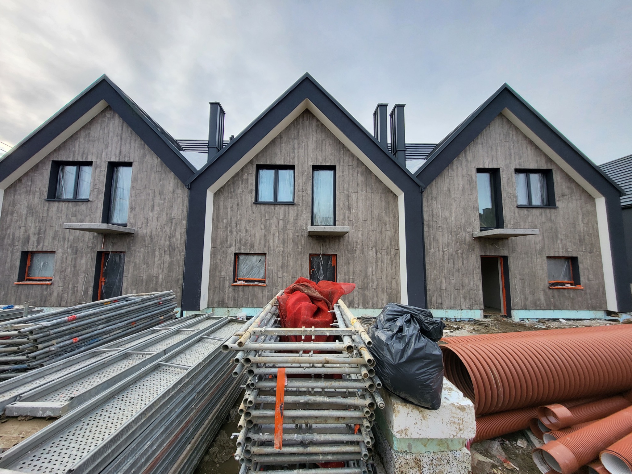 Nowoczesne domy w zabudowie szeregowej z szarą elewacją imitującą drewno, widoczne rusztowania i materiały budowlane, stan surowy otwarty.