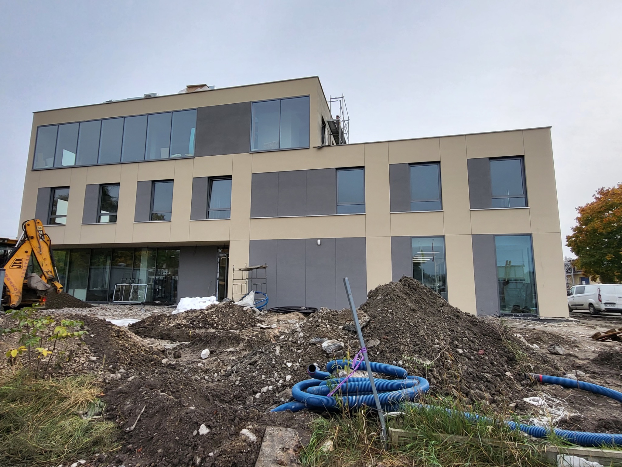 Dwukondygnacyjny budynek w trakcie prac elewacyjnych, z ziemią, rurami i koparką na pierwszym planie, oraz rusztowaniem przy ścianie. Budynek z dużymi oknami i panelami.