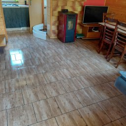 Ekipa czystości - Ukończona podłoga z płytek imitujących drewno w jasnym salonie z drewnianymi elementami, piecem i schodami. Widoczne drobne elementy wyposażenia.