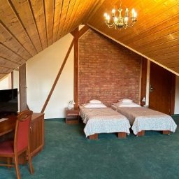 Ekipa czystości - Pokój na poddaszu z dwoma łóżkami, biurkiem i ceglaną ścianą. Drewniane wykończenie sufitu i zielona wykładzina. Widoczny żyrandol i lampka nocna.