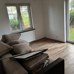 Ekipa czystości - Pusty pokój z brązową podłogą i kanapą z poduszkami, na której leżą obrazy w ramkach. Widok na ogród przez okno. Przygotowanie do sprzątania po remoncie.