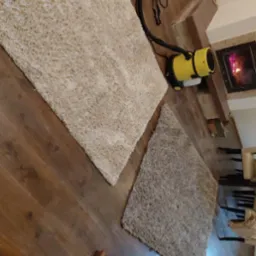 Dwa dywany o różnej fakturze i kolorze na drewnianej podłodze, obok żółty odkurzacz piorący i kominek w tle. Widok z góry, ukośny kadr.