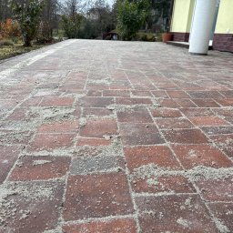 Ekipa czystości - Czerwona kostka brukowa z piaskiem na powierzchni, widok z perspektywy chodnika, fragment budynku w tle, zieleń po bokach. Kostka przygotowana do czyszczenia.