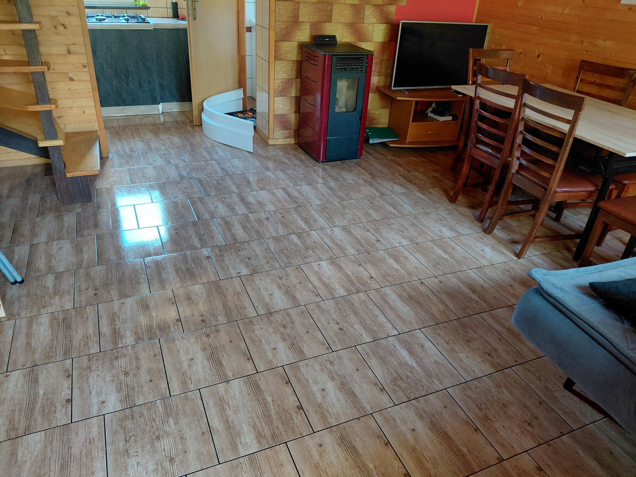 Ukończona podłoga z płytek imitujących drewno w jasnym salonie z drewnianymi elementami, piecem i schodami. Widoczne drobne elementy wyposażenia.