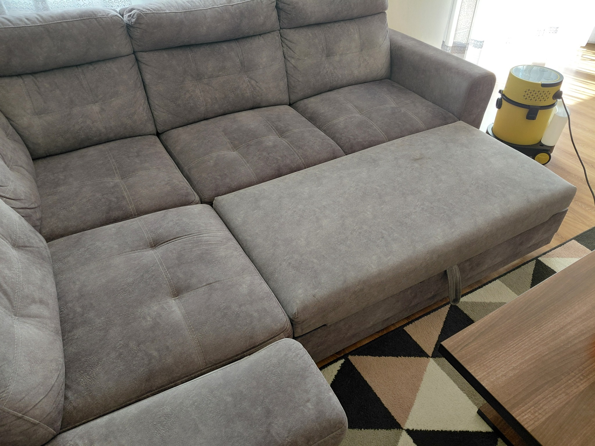 Szara, rozłożona sofa z nakładką i odkurzaczem piorącym obok. Widoczny fragment stolika kawowego i dywanu w geometryczne wzory.