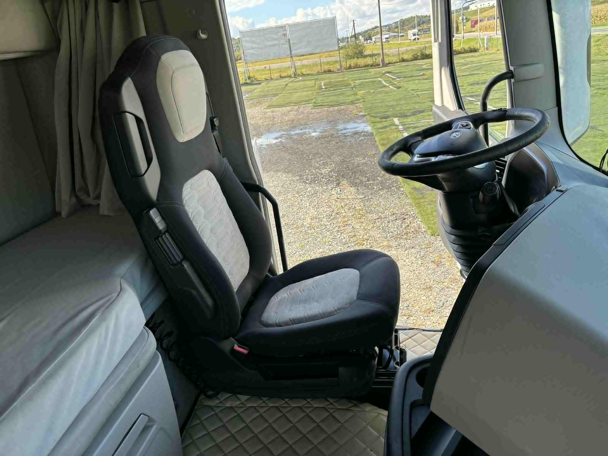 Wnętrze kabiny ciężarówki: fotel kierowcy z szaro-czarną tapicerką w geometryczny wzór, widok na kierownicę i otoczenie za oknem w słoneczny dzień.