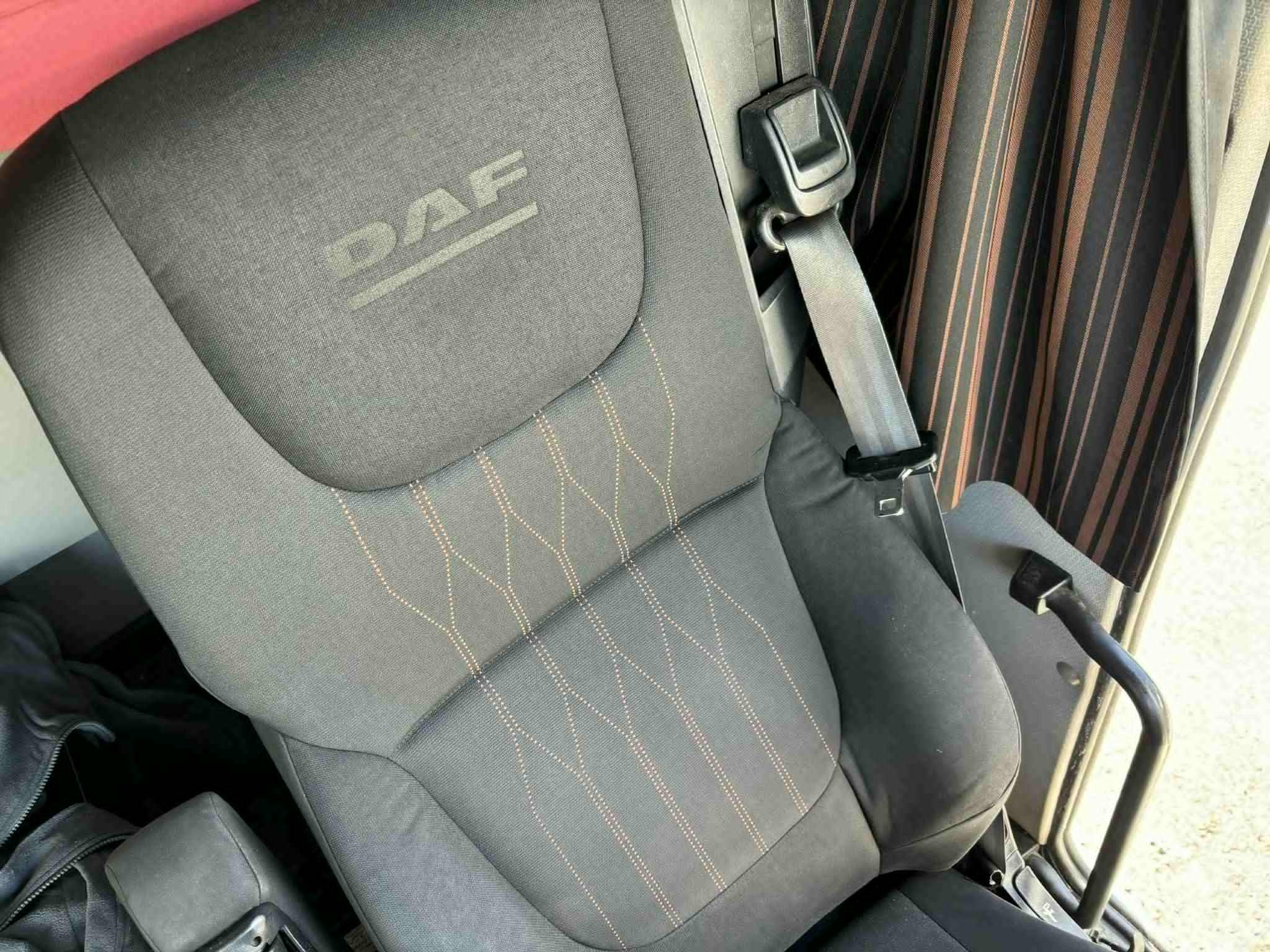 Szary fotel kierowcy DAF z pasami bezpieczeństwa i pomarańczowymi przeszyciami, widok z boku, wnętrze kabiny pojazdu z zasłonami w tle.