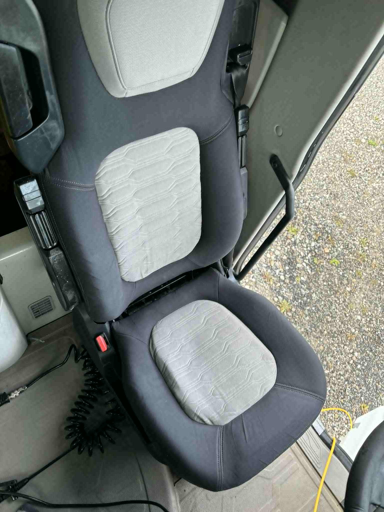 Wnętrze kabiny pojazdu z ciemnoszarym fotelem z jasnoszarą tapicerką, widoczny pas bezpieczeństwa i elementy sterowania. Fotel czysty i zadbany.