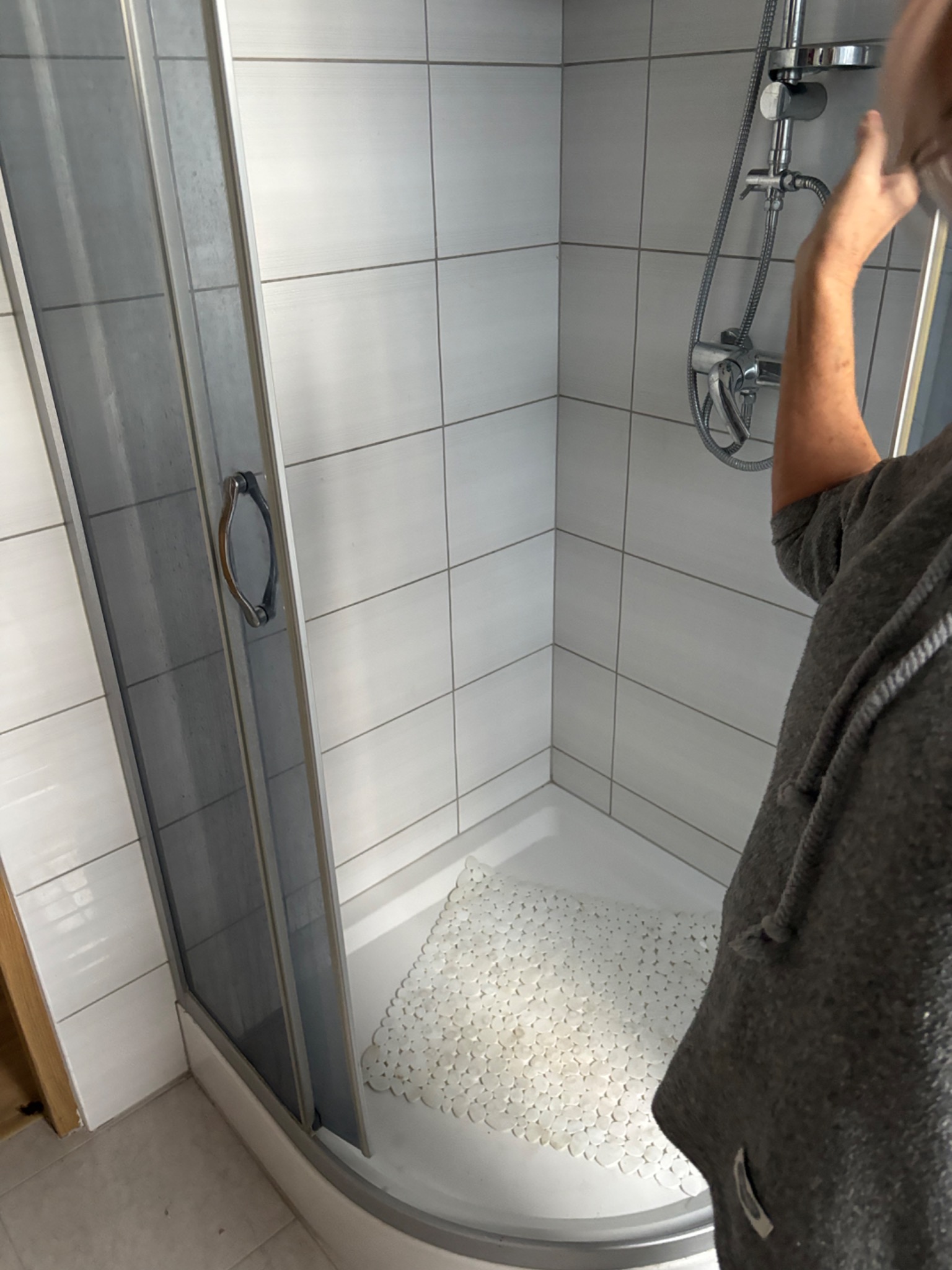 Błyszcząca kabina prysznicowa z chromowaną armaturą i białymi płytkami, widok z boku, osoba w szarej bluzie częściowo zasłania kadr. Czysta mata antypoślizgowa w brodziku.