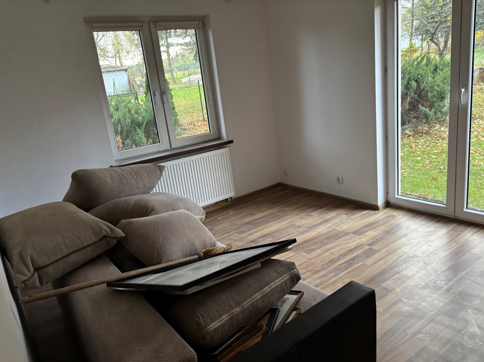 Pusty pokój z brązową podłogą i kanapą z poduszkami, na której leżą obrazy w ramkach. Widok na ogród przez okno. Przygotowanie do sprzątania po remoncie.