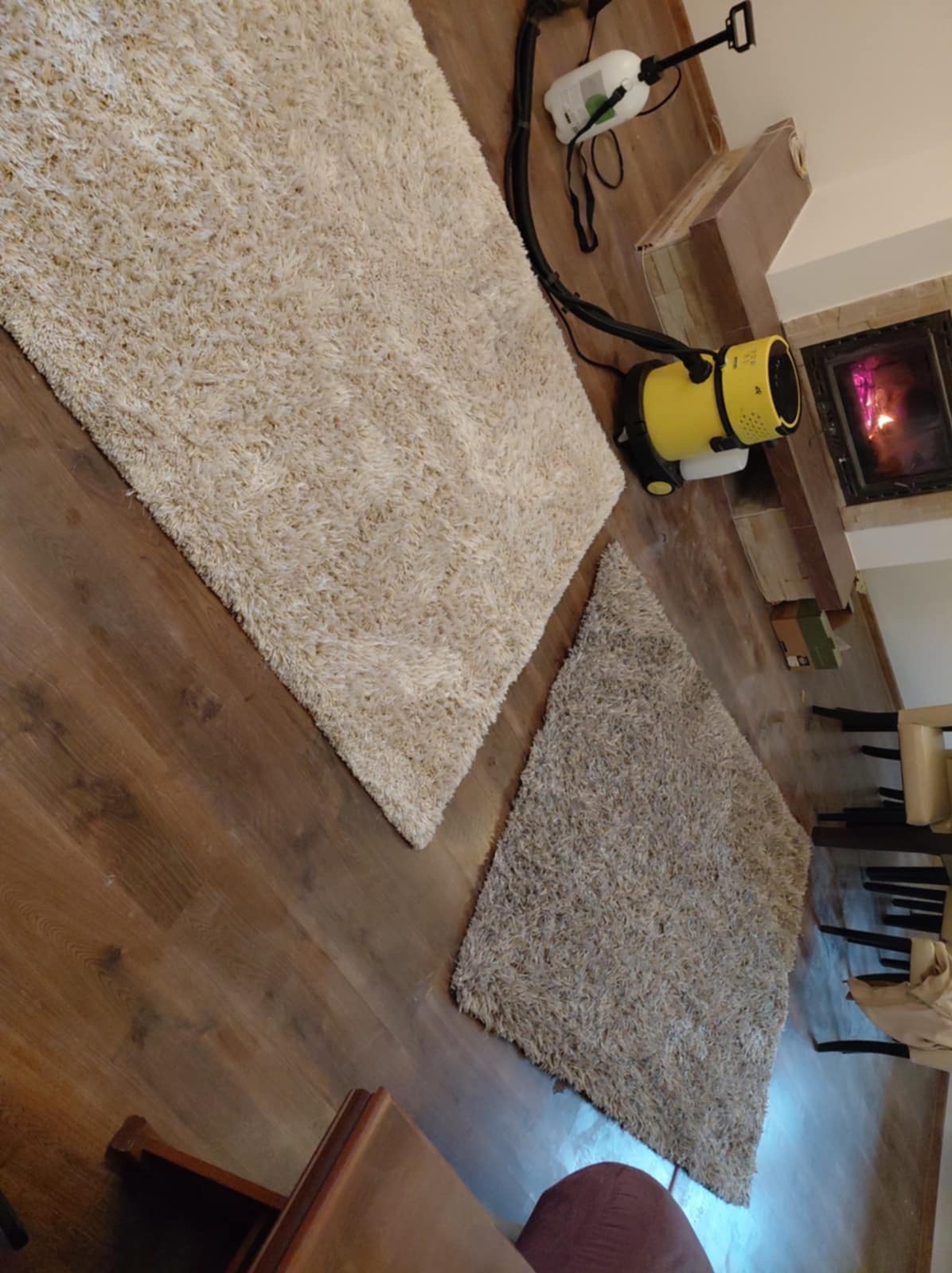 Dwa dywany o różnej fakturze i kolorze na drewnianej podłodze, obok żółty odkurzacz piorący i kominek w tle. Widok z góry, ukośny kadr.