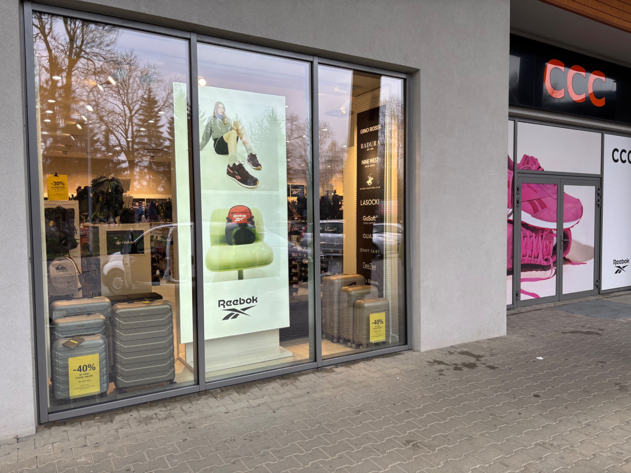 Witryna sklepowa z walizkami i obuwiem sportowym marki Reebok, obok widoczny sklep CCC. Na szybie naklejki z promocjami -30% i -40%.