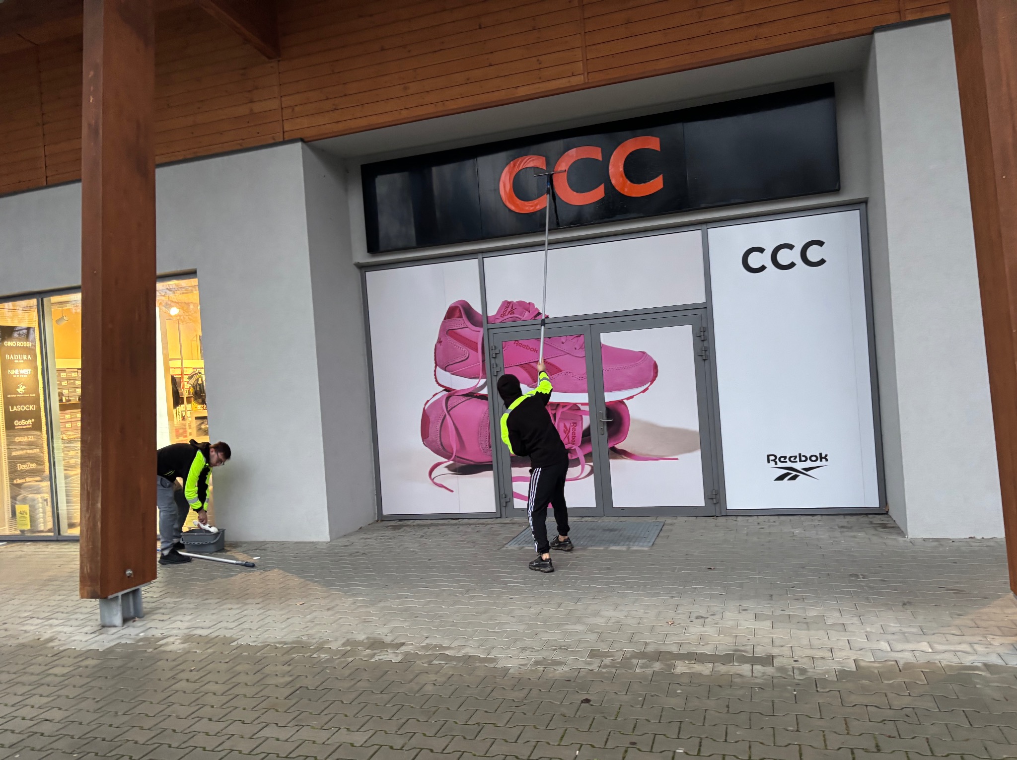 Pracownicy w odblaskowych kamizelkach myją okna i witryny sklepu obuwniczego CCC. Jeden z nich używa mopa na długim kiju, drugi czyści dolne partie. Widoczny szyld i reklama butów.