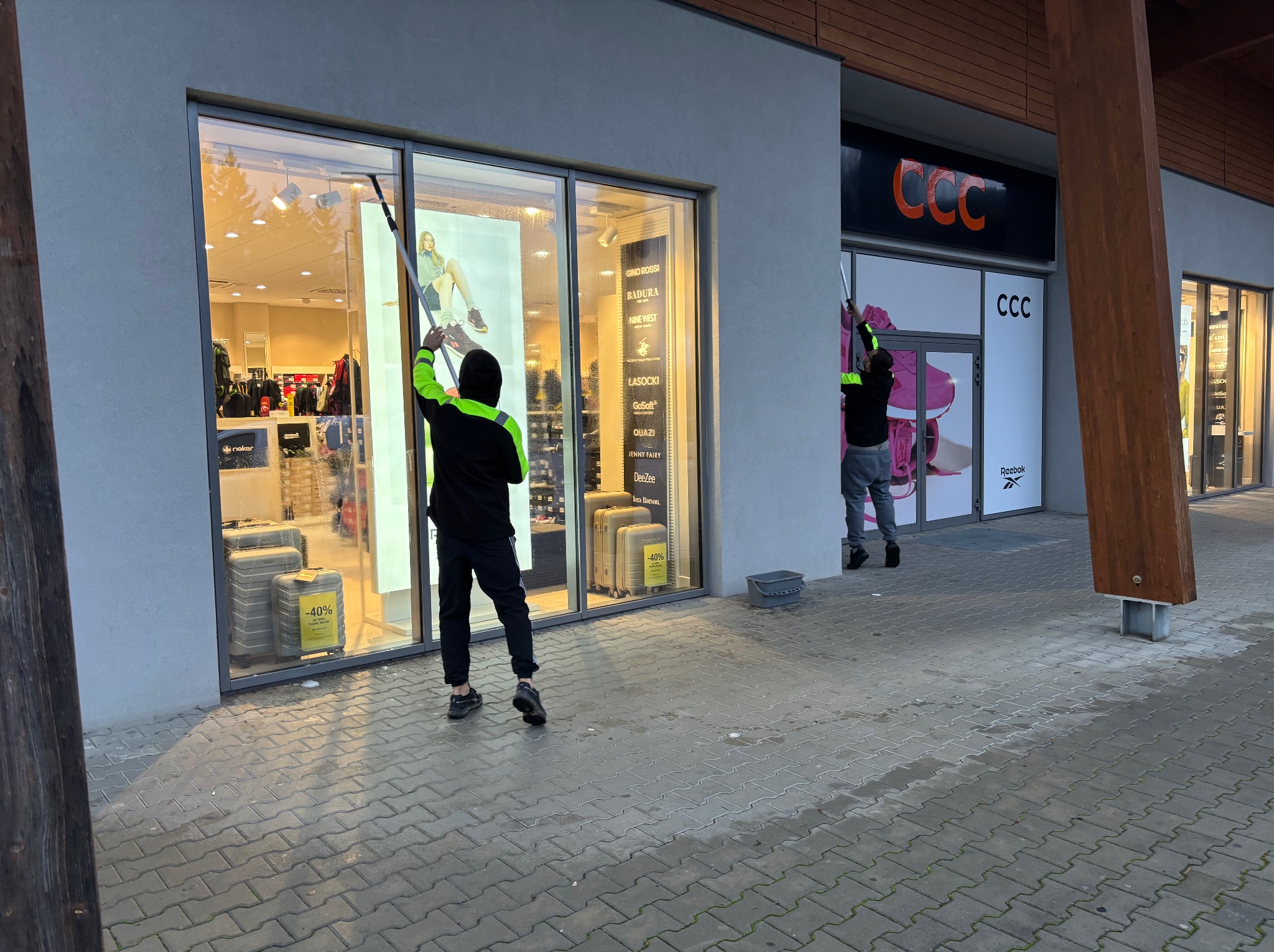 Pracownicy w odblaskowych kurtkach myją okna wystawowe sklepów w pasażu handlowym. Widoczne marki: CCC, Gino Rossi, Badura, Lasocki, Reebok. Na oknach promocje -40%.