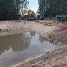 Żółta koparka przy wykopanym stawie z wodą, na tle drzew i nieba. Widoczny niebieski samochód ciężarowy. Brzegi stawu z ziemi i piasku.