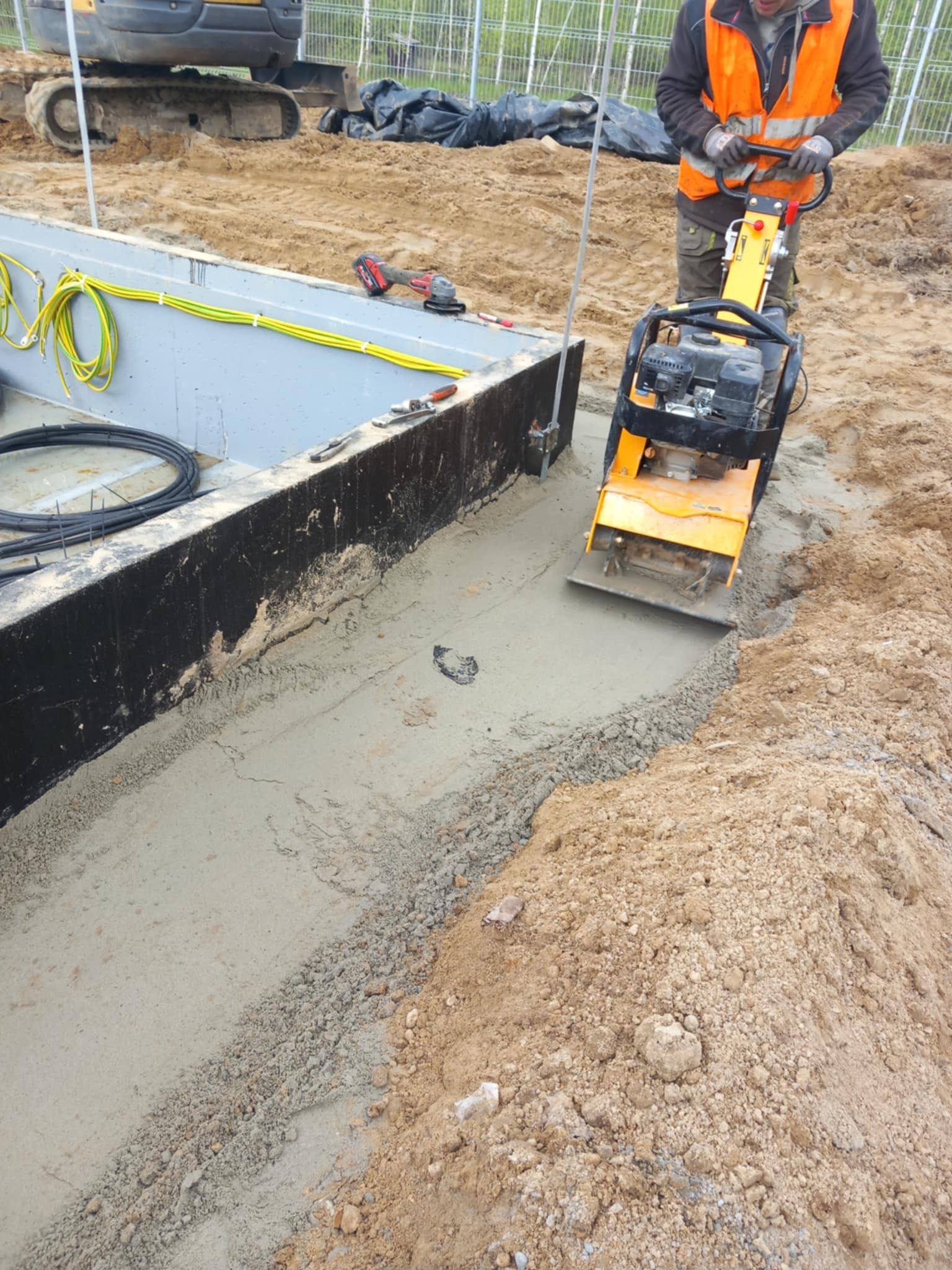 Pracownik zagęszcza podłoże ubijarką mechaniczną przy betonowym fundamencie z okablowaniem. W tle koparka i ogrodzenie.