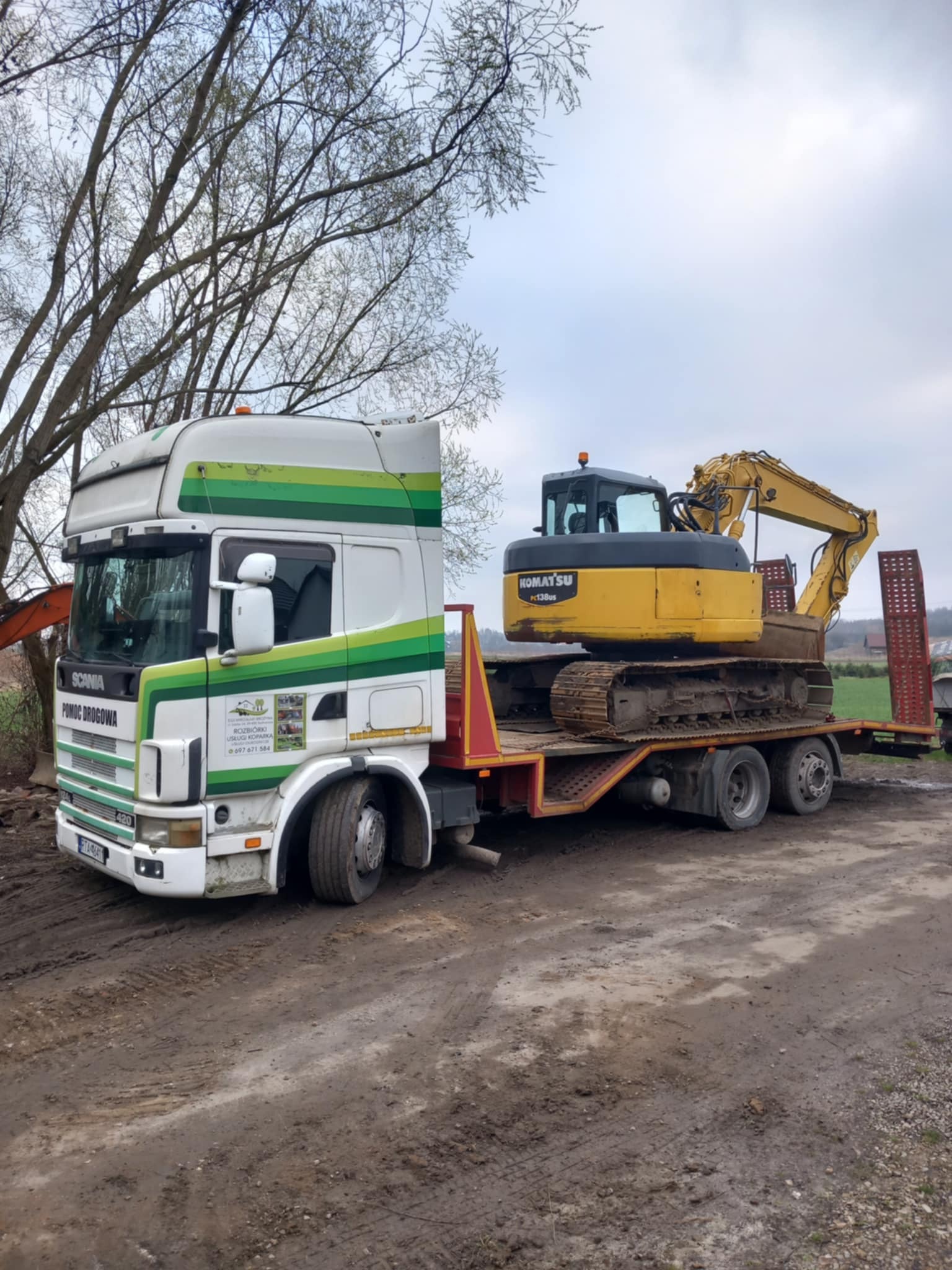 Ciężarówka Scania z żółtą koparką Komatsu na lawecie, teren błotnisty, drzewa w tle. Koparka gotowa do pracy.