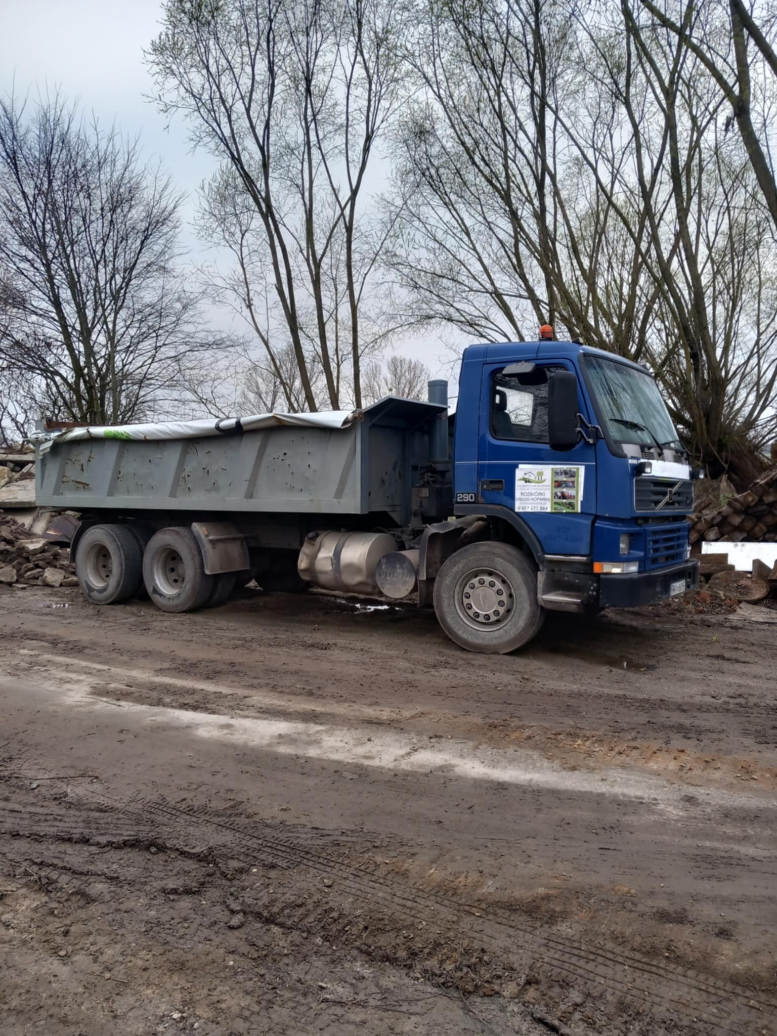 Niebieska ciężarówka Volvo 290 z przykrytą plandeką skrzynią ładunkową stoi na błotnistym terenie na tle drzew bez liści i gruzu. Widoczne logo firmy na kabinie.