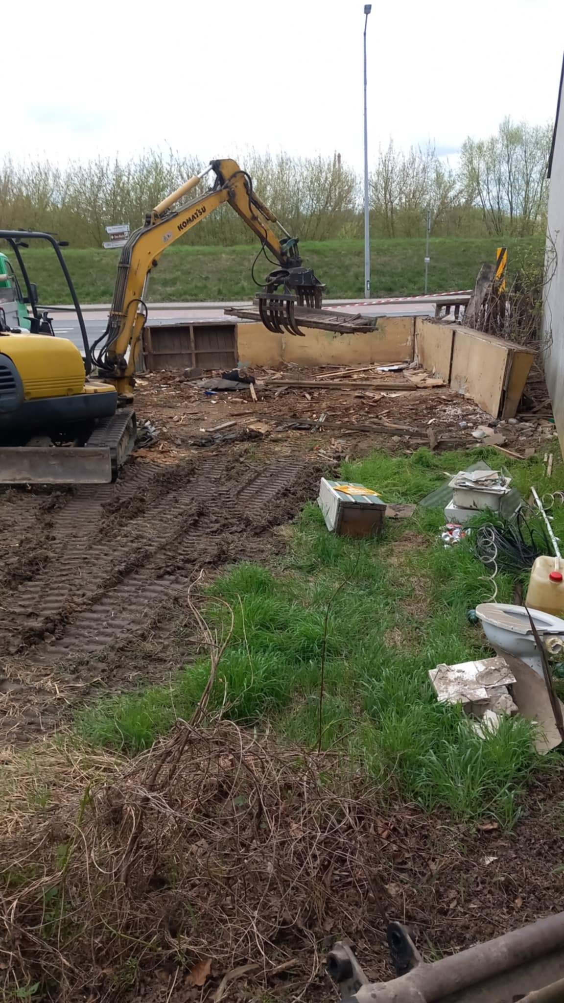Żółta koparka Komatsu z chwytakiem podczas rozbiórki fundamentów budynku. W tle widać zieleń i drogę.