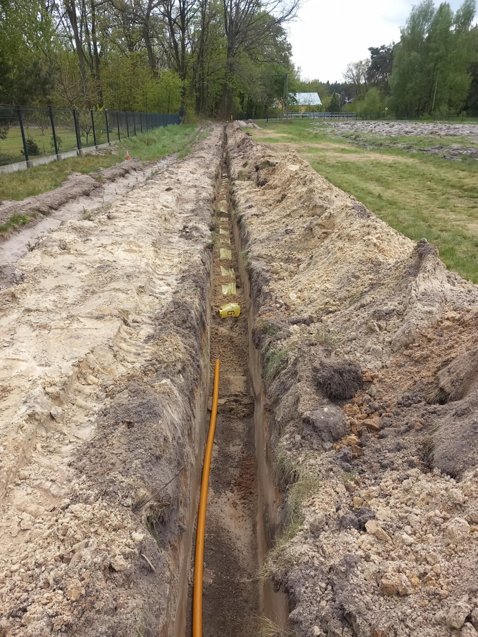Wykop pod instalacje w Suchorzowie z widoczną pomarańczową rurą i żółtą taśmą ostrzegawczą. Wykop prowadzony wzdłuż ogrodzenia na terenie zielonym.