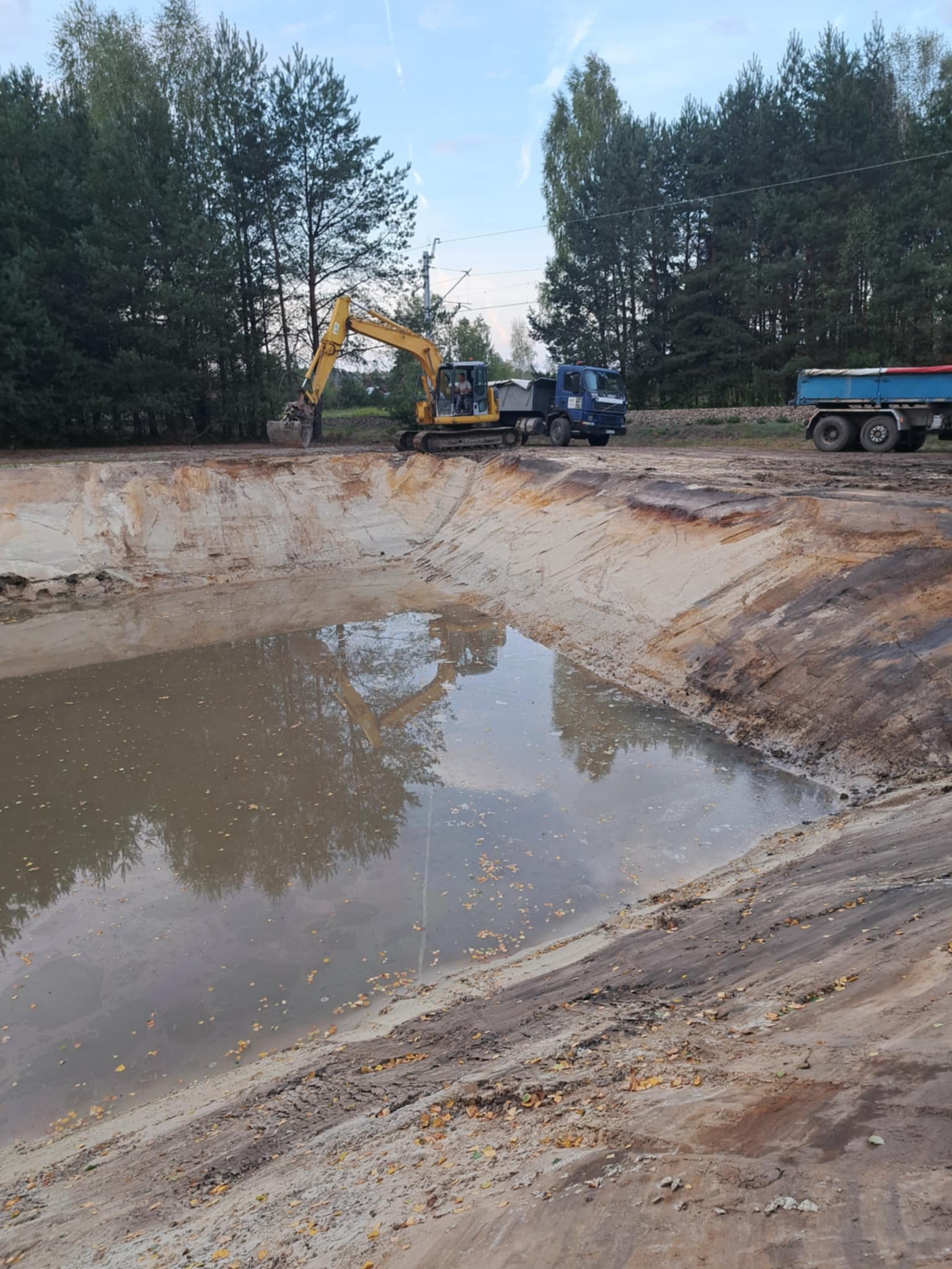 Żółta koparka przy wykopanym stawie z wodą, na tle drzew i nieba. Widoczny niebieski samochód ciężarowy. Brzegi stawu z ziemi i piasku.