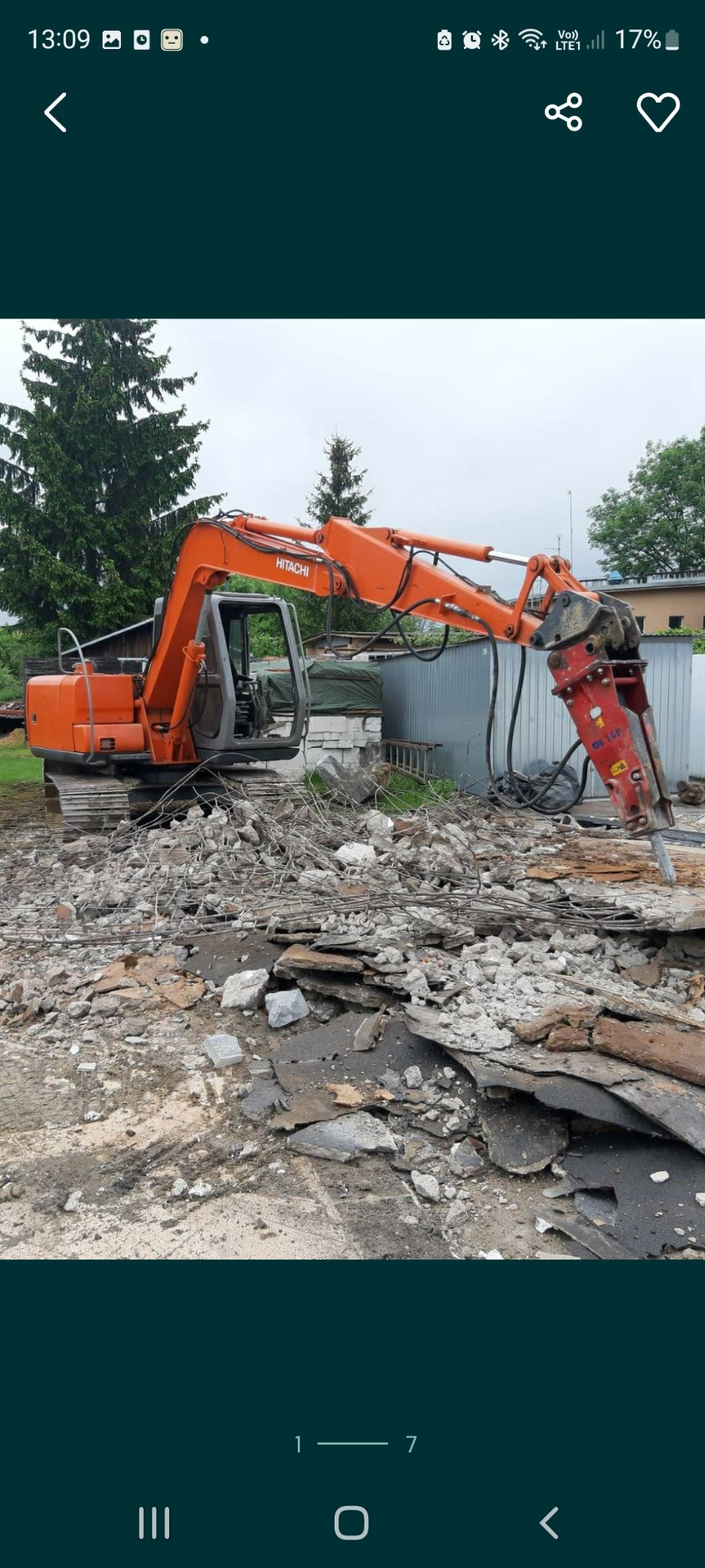 Pomarańczowy młot hydrauliczny Hitachi rozbija beton na placu budowy. Widoczne gruz i elementy zbrojenia. W tle zieleń drzew i ogrodzenie.