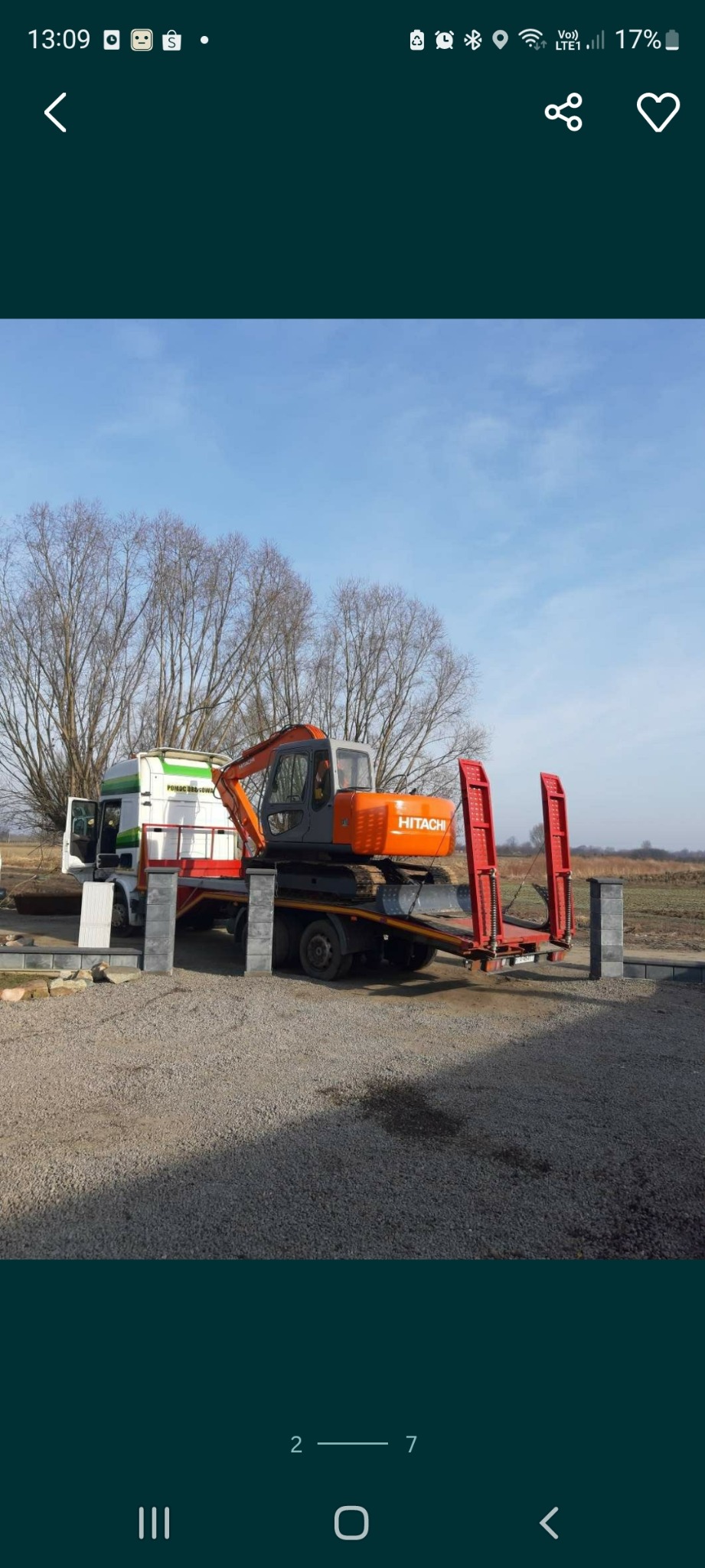 Pomarańczowa koparka Hitachi na lawecie ciężarówki, widoczna na żwirowym terenie z drzewami w tle. Laweta z czerwonymi elementami, obok szare słupki.