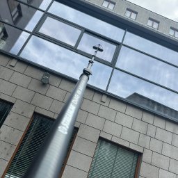 AlpinPeak - Mycie okien na wysokości budynku biurowego za pomocą teleskopowej lancy ze szczotką. Widoczny fragment elewacji z oknami i odbicie nieba w szybach.