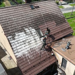 AlpinPeak - Częściowo umyty dach z brązowej dachówki. Dwóch pracowników w uprzężach czyści dach myjką ciśnieniową. Widok z góry na dom z ogrodem i ulicą w tle.