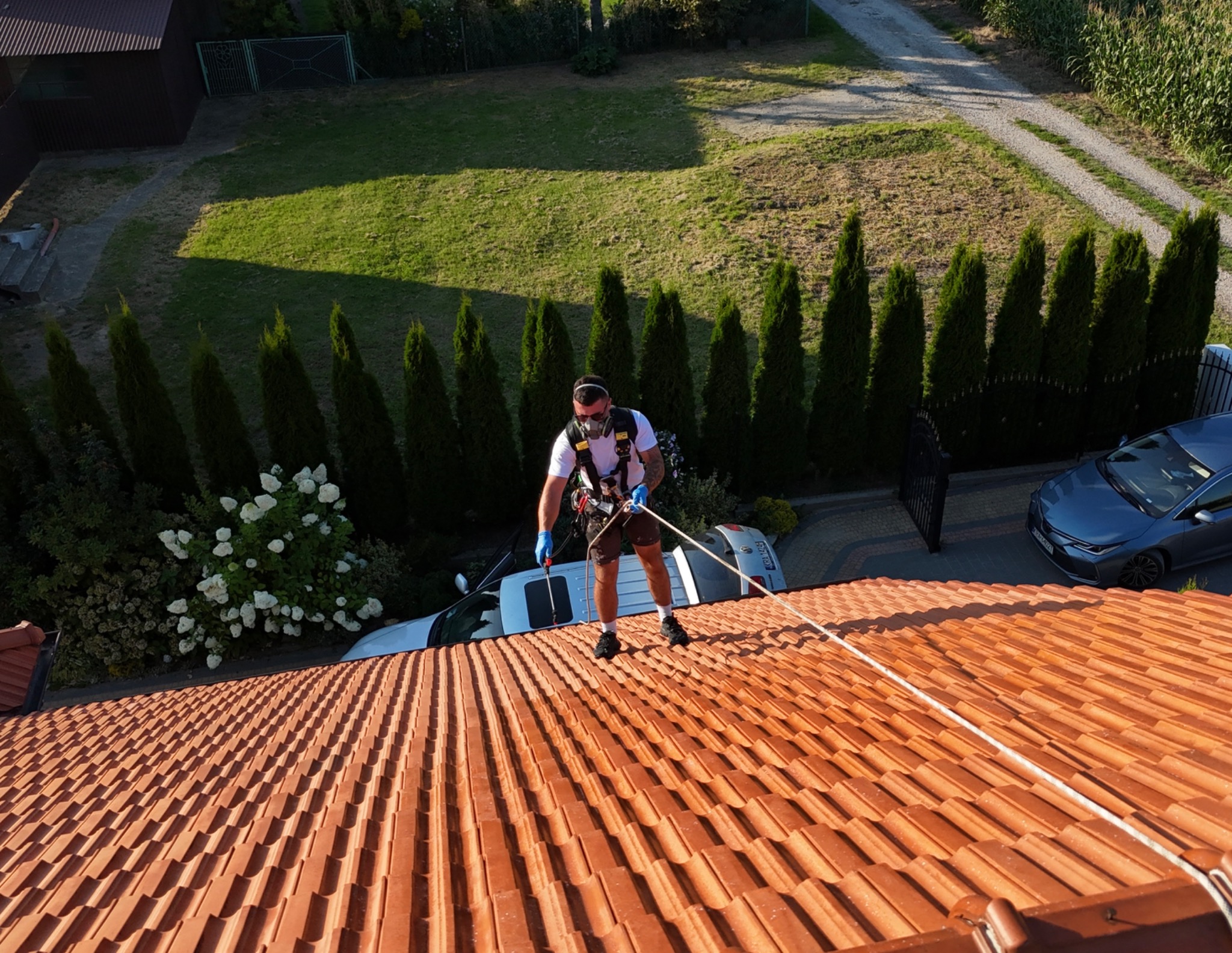 Pracownik w uprzęży na dachu z czerwonej dachówki, myje go lancą ciśnieniową. W tle ogród, samochody i budynki. Usługa mycia dachów.