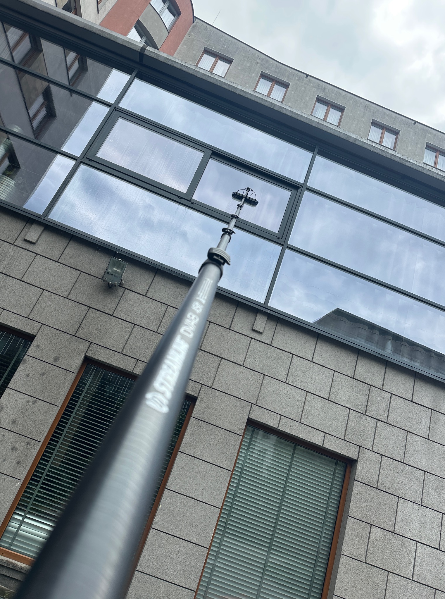 Mycie okien na wysokości budynku biurowego za pomocą teleskopowej lancy ze szczotką. Widoczny fragment elewacji z oknami i odbicie nieba w szybach.