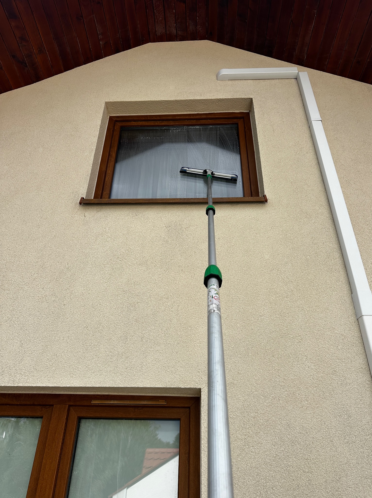 Mycie okna na wysokości za pomocą teleskopowego kija z myjką na tle elewacji domu i brązowej stolarki okiennej.
