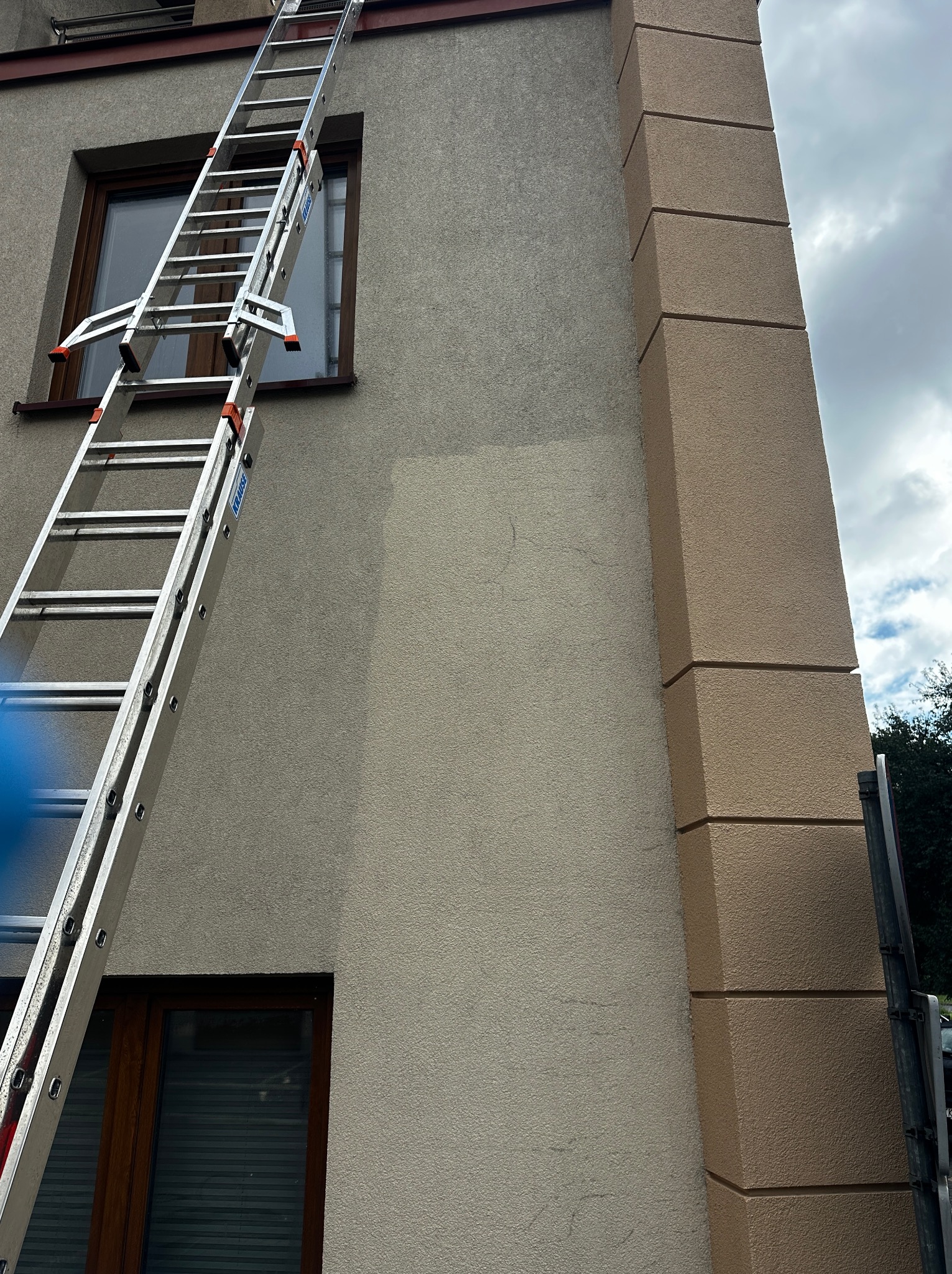 Długa, aluminiowa drabina oparta o elewację budynku z oknami. Widoczne fragmenty elewacji i okna. Pochmurne niebo w tle.