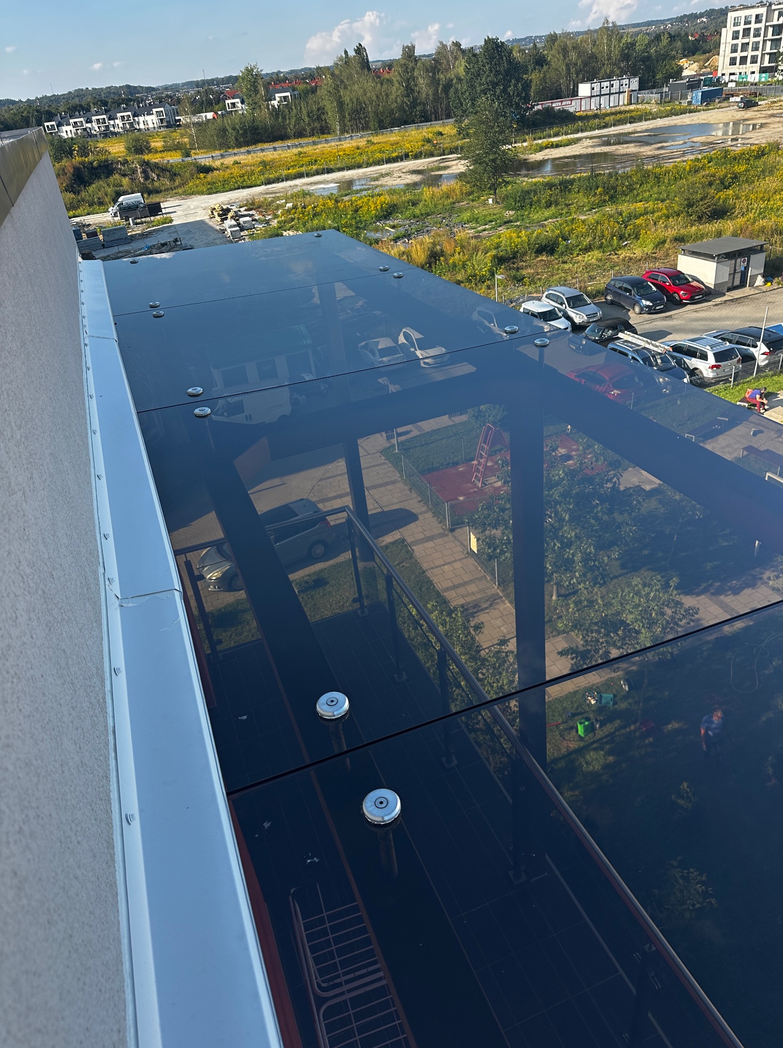 Szklane zadaszenie nad balkonem z widokiem na okolicę, zabezpieczone metalowymi śrubami. Widoczne budynki mieszkalne i zieleń w tle.