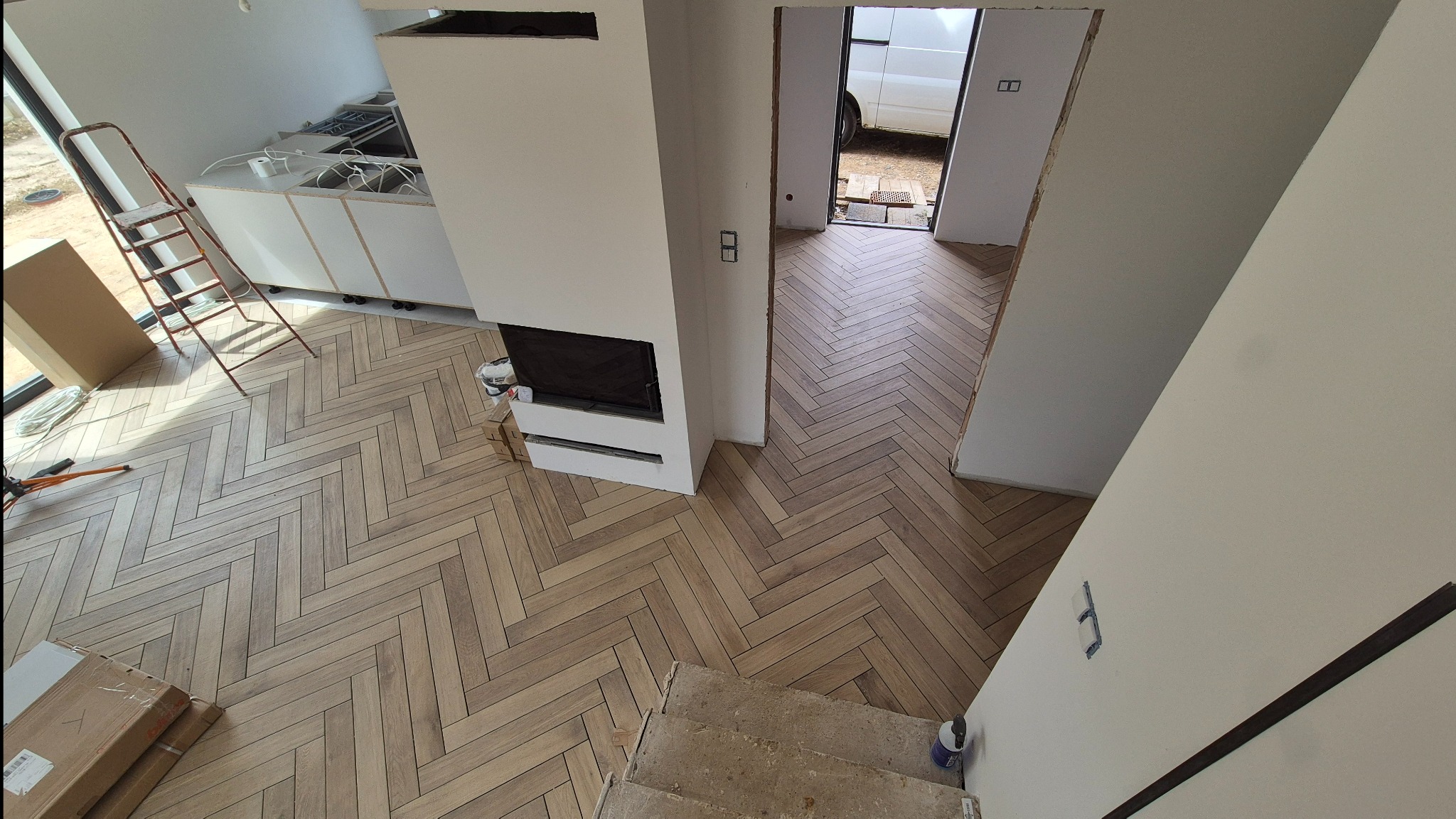 Jasna, drewnopodobna podłoga ułożona w jodełkę francuską w nowym wnętrzu. Widoczny fragment schodów, biała zabudowa kuchenna i kominek. W tle otwarte drzwi.