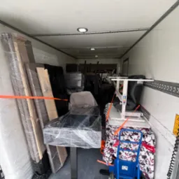 Wnętrze busa wypełnione meblami i sprzętami zabezpieczonymi folią stretch i pasami transportowymi, gotowymi do przeprowadzki. Widoczny wózek transportowy.
