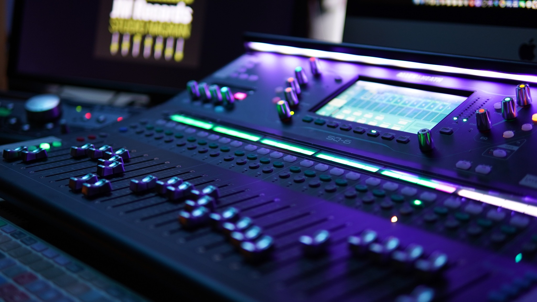 Profesjonalny mikser audio SQ-6 z podświetleniem LED w odcieniach fioletu i zieleni. Widoczne suwaki, pokrętła i ekran. W tle monitory.