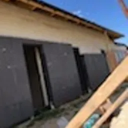 Ocieplanie ściany budynku płytami izolacyjnymi z widoczną konstrukcją dachu i niebem w tle. Prace w toku, jasna elewacja, brązowe płyty izolacyjne.