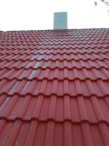 Czerwona dachówka ceramiczna na skośnym dachu domu jednorodzinnego, widoczny komin z blachy w tle. Nowe pokrycie dachu.