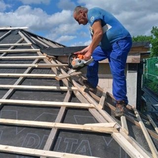 Dekarz tnie pilarką drewniane łaty na dachu pokrytym czarną membraną. Widoczne niebo i fragmenty zieleni w tle, praca w toku.