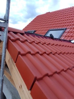 Czerwony dach z dachówki ceramicznej z oknami połaciowymi, widok z dołu. Fragment rusztowania po lewej. Niebo częściowo zachmurzone.