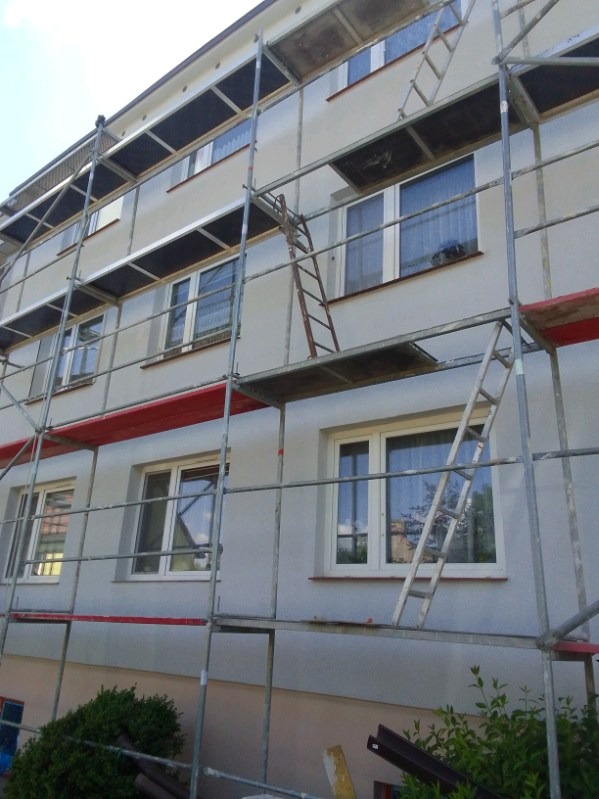 Elewacja budynku w trakcie szpachlowania, widoczne rusztowania i drabiny. Świeżo nałożona szpachla na ścianie. Częściowo widoczne okna.