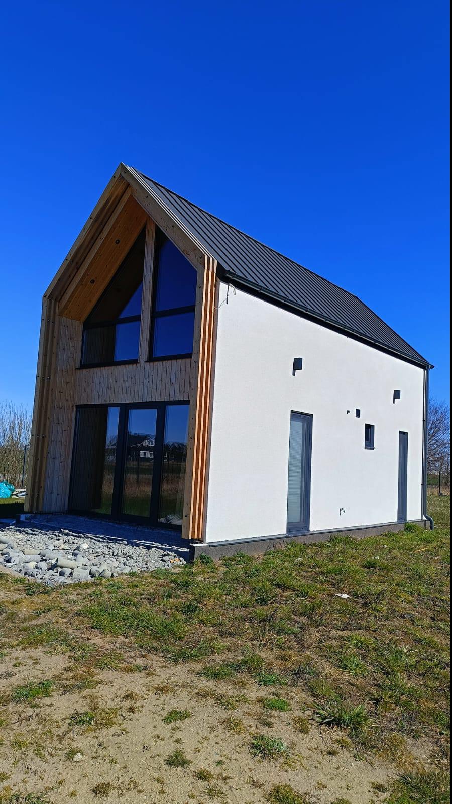 Nowoczesny dom z białymi ścianami i drewnianą fasadą pod ciemnym dachem na tle błękitnego nieba. Widoczne okna i drzwi w ciemnych ramach.