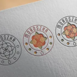Trzy okrągłe, tłoczone logotypy 'Moroszka' na szarym papierze o subtelnej fakturze. Dwa logotypy z kolorowym wypełnieniem, jeden konturowy. Minimalistyczny design.