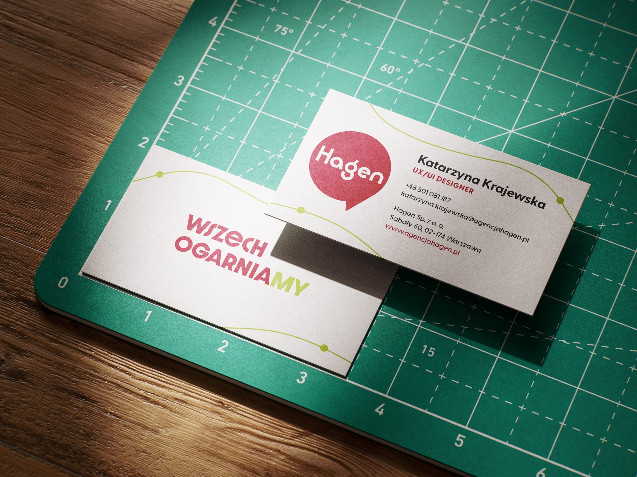 Dwie wizytówki firmy Hagen na zielonej macie do cięcia. Jedna z logo i hasłem 'Wszech Ogarniamy', druga z danymi kontaktowymi UX/UI Designera.