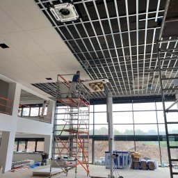 Sławomir Kamiński - Montaż sufitu podwieszanego w biurze: pracownik na rusztowaniu instaluje panele sufitowe w metalowej konstrukcji. Widoczne okna i materiały budowlane.